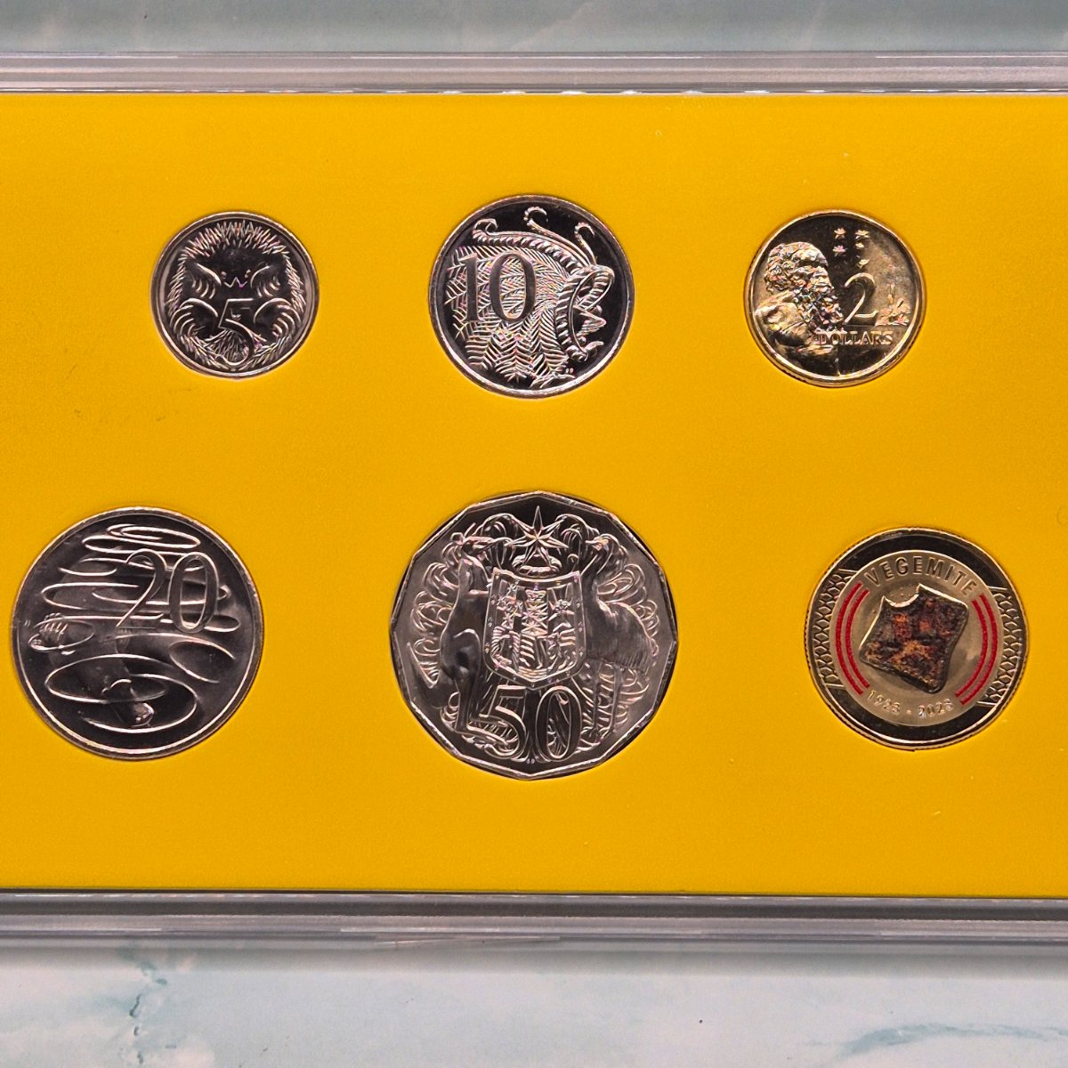 Set of six 2023 Royal Australian Mint Vegemite Year set coins including coloured $1 Vegemite on Toast in a display case with a yellow background - reverse side