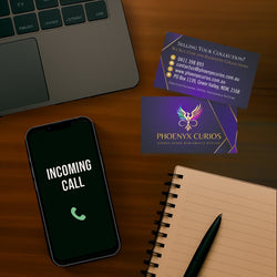 Phone with 'Incoming Call' display, notebook, pen, and two business cards on a wooden surface. Contact us at Phoenyx Curios